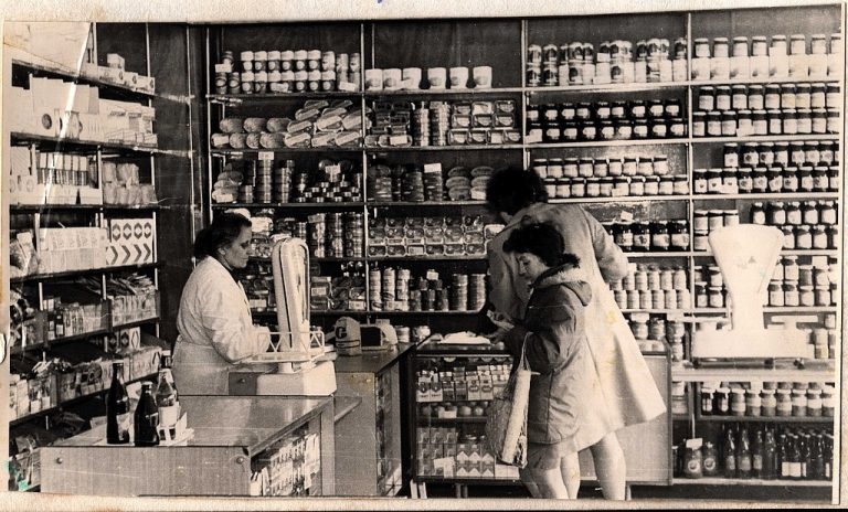 Fotografia czarno-biała przedstawiająca wnętrze sklepu delikatesowego oraz klientów w latach 80.