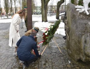 Dwóch mężczyzn stawiających zapalone znicze pod wieńcem, nad nimi pochylona kobieta.