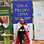 Na zdjęciu widać pana w garniturze i białej szacie z czerwonym krzyżem. Za templariuszem stoją rolapy Biblioteki Publicznej w Gołdapi, ,,Cała Polska czyta dzieciom’’ oraz patronat honorowy burmistrza Gołdapi.