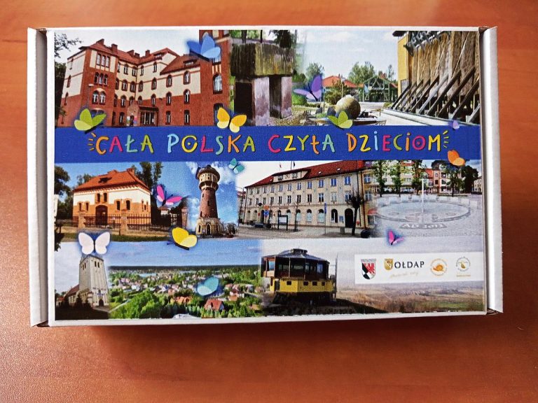 Zdjęcie przedstawia pudełko puzzli z akcji ,,Cała Polska czyta dzieciom’’ . Na pudełku widnieje obrazek Biblioteki Publicznej w Gołdapi, tężni, Muzeum w Gołdapi, Urzędu Miasta Wieży ciśnień.