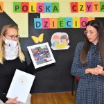 pani dyrektor Biblioteki Publicznej w Gołdapi trzymająca w ręku puzzle w ramach akcji „Cała Polska czyta dzieciom”, w drugiej zaś białą teczkę z logo biblioteki, obok stoi pani Skarbnik