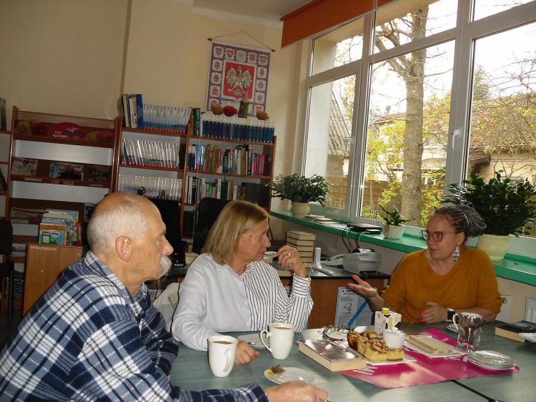 Dwie kobiety i mężczyzna przy zielonym stole rozmawiają o przeczytanej książce. Na stole jest widoczny plakat DKK.