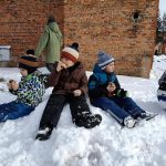 Dzieci ubrane ciepło siedzą na śniegu i jedzą kiełbaski.