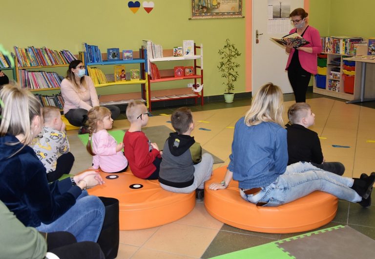 Dzieci siedzą na pufach praz pani w maseczce czyta książkę.