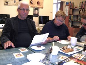 Zdjęcie przedstawia dwie kobiety i mężczyznę trzymającego w dłoni kartkę (odczytuje recenzję książki), na stole widać słodkości, białe kubki i logotyp DKK.