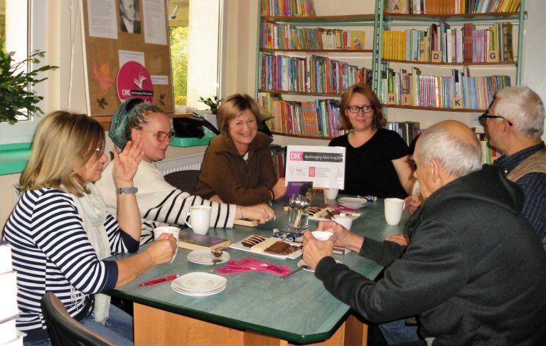 Przy zielonym stole siedzą cztery kobiety i dwóch mężczyzn(uczestnicy DKK). Na stole widać logotyp, zakładki DKK , białą zastawę i słodkości.