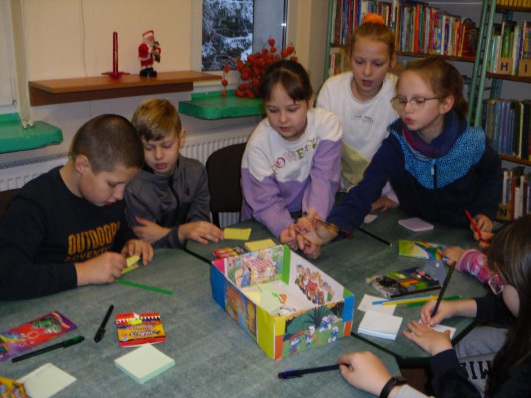 Na zdjęciu widać dzieci, które biorą udział w zabawie, na stole leżą kredki, karteczki, stoi kolorowe pudełko.