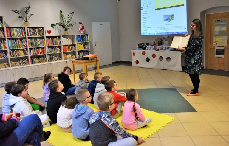 Grupa dzieci siedzi na piankowych matach w wypożyczalni dla dzieci, skierowana w stronę stojącej bibliotekarki, która pokazuje im księgę inwentarzową.