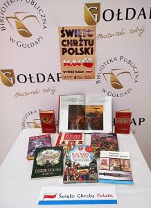 Książki na temat historii Polski prezentowane na stole przykrytym obrusem. Nad stołem na ściance z ologowaniami biblioteki plakat informujący o rocznicy Chrztu Polski.