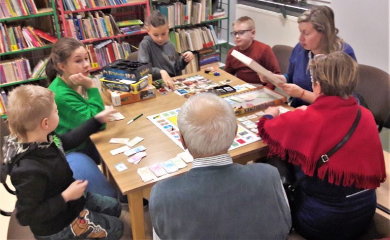 Grupka dzieci i dorosłych siedząca dookoła stołów biblioteki gra w gry planszowe.