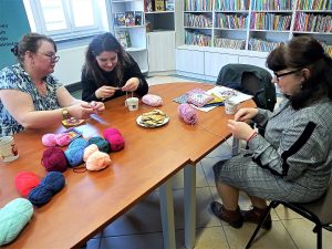 3 kobiety siedzące przy stołach w wypożyczalni książek, wykonują pracę na szydełku.