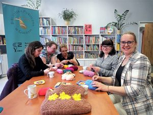 5 kobiet siedzących przy stołach w wypożyczalni książek, wykonują pracę na szydełku.