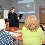 Grupa szkoleniowa widziana od tyłu, siedzi przy stołach i patrzy w stronę prowadzącej szkolenie, która pokazuje coś na tablecie. W tle na ścianie slajd prezentacji multimedialnej.