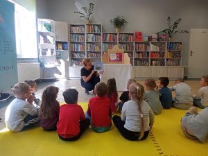 Pani pokazuje siedzącym w wypożyczalni biblioteki dzieciom książkę.