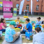 Grupa dzieci siedzi na piankach przed budynkiem biblioteki, trzymają w dłoniach książki.