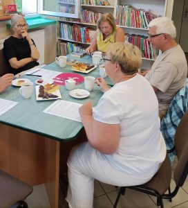 2. Cztery osoby siedzą przy stoliku na którym znajduje się ciasto i kubki z kawą i herbata. Uczestnicy spotkania rozmawiają ze sobą.