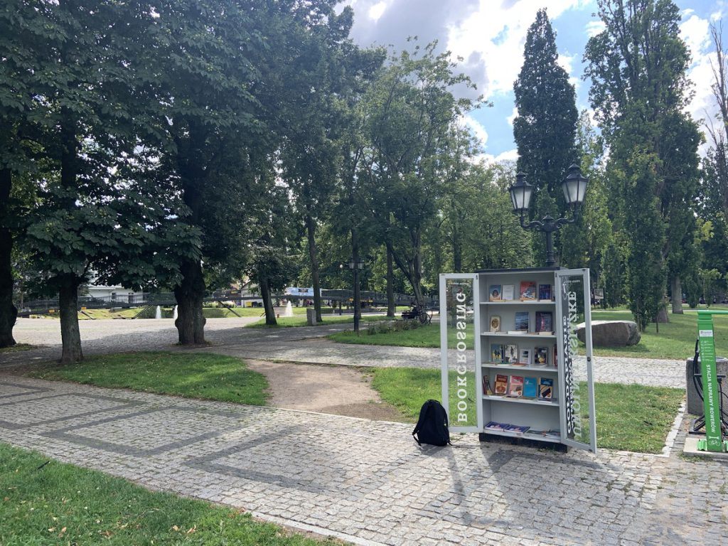 otwarta szafa bookcrossingowa z książkami na tle parku miejskiego w Gołdapi