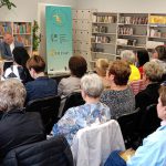 Bibliotekarka i ksiądz siedzą przy stoliku w wypożyczalni książek przed publicznością spotkania autorskiego.