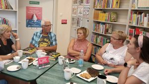 Pięć osób siedzą przy stoliku na którym znajduje się ciasto, kubki z kawą i herbata. Na stole znajduje się również książka, która jest tematem rozmów uczestników.