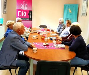 Klubowicze Dyskusyjnego Klubu Książki siedzący za stołem w tle widać rolap DKK i biblioteki.