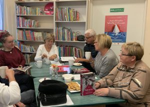 Grupa osób siedzi w bibliotece przy stoliku i rozmawiają ze sobą. W rękach trzymają książki lub popijają herbatkę.