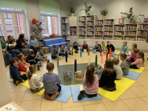 Grupa dzieci siedzi w koło na piankach. W tle regały z książkami.