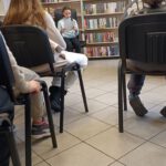 Grupa uczniów siedzi w bibliotece, jedna dziewczynka czyta uczniom