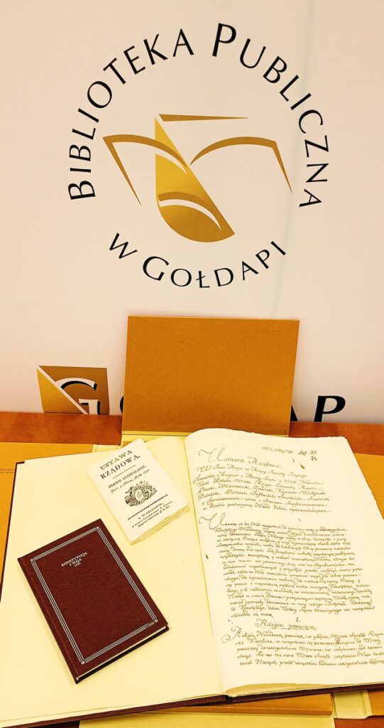 Na fotografii na pierwszym planie widnieje otwarta książka wraz z małymi książeczkami, w tle logo biblioteki.