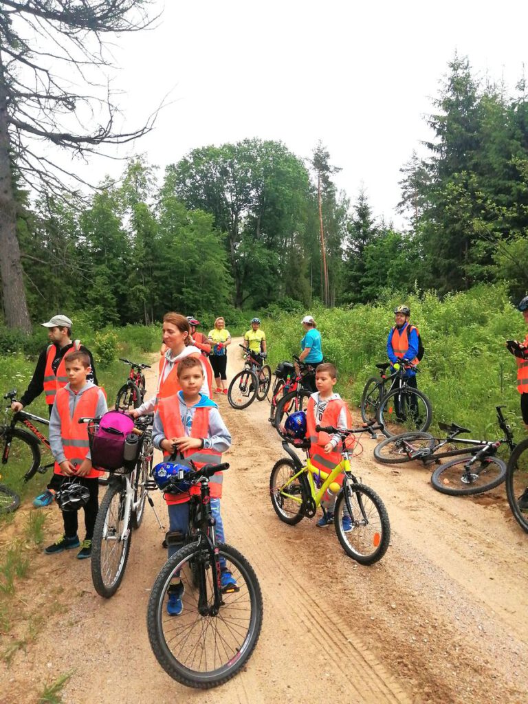 Uczestnicy rajdu rowerowego w kamizelkach odblaskowych na leśnej drodze, wśród zieleni.