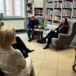 Poetka i prowadzący spotkanie siedzą w szarych fotelach przy okrągłym stoliku zwróceni w stronę siedzącej w pierwszym rzędzie publiczności. Po prawej stronie na małym okrągłym stoliku ustawione 3 książki autorki.