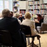 Poetka i prowadzący spotkanie siedzą w fotelach zwróceni w stronę publiczności. Po prawej stronie stoi telewizor ze zdjęciem autorki na ekranie.