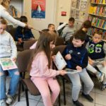 Grupa dzieci siedzi na krzesłach i czyta książki. W tle widać regały z książkami.