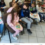 Grupa dzieci siedzi na krzesłach i czyta książki. W tle widać regały z książkami.