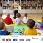 Grupa przedszkolaków siedzi na piankach przed nimi ustawiono stolik z teatrzykiem, który czyta kobieta.