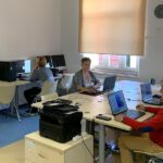 Grupa uczestników warsztatów komputerowych 50+ pracuje przy komputerach stacjonarnych i laptopach w sali szkoleniowej Biblioteki Publicznej w Gołdapi.