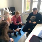 Grupka starszych osób w pracowni komputerowej trzymają w rękach komórki. Wymieniają się informacjami na temat obsługi.