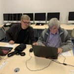 Dwóch mężczyzn pracuje razem przy laptopie, jeden z nich pomaga drugiemu przy obsłudze komputera.