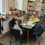 Dwie panie tłumaczą dzieciom, które znajdują się w bibliotece.