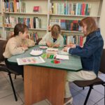 Pani w dżinsowej kurtce siedzi z dwiema dziewczynkami i układają kolorowe koraliki. W tle widać regały z książkami.