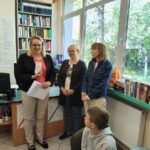 Trzy panie stoją przy oknie, przed nimi dzieci. Jedna z pań pokazuje dzieciom kartę biblioteczną.