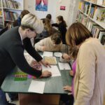 Dwie panie tłumaczą dzieciom, które siedzą przy stolikach w bibliotece.