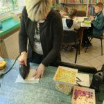 Pani w jasnych włosach prasuje kolorowe koraliki. W tle widać dzieci układające koraliki oraz regały z książkami.
