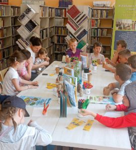 Fotografia przedstawia dzieci siedzące przy stole na którym są poustawiane książki i przybory potrzebne do twórczej pracy.