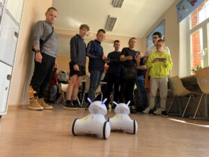 Grupa młodzieży wraz z opiekunką stoją przed robotami. Dwoje chłopców trzyma tablety w dłoniach którymi sterują roboty.