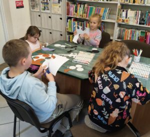 Czworo dzieci wycina z kolorowego papieru, siedzą przy stole na którym leżą mazaki i kartki.
