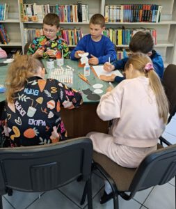 Pięcioro dzieci wycina z kolorowego papieru, siedzą przy stole na którym leżą mazaki i kartki.