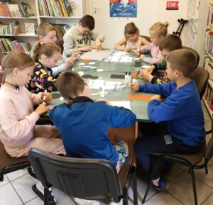Dzieci wycina z kolorowego papieru, siedzą przy stole na którym leżą mazaki i kartki.