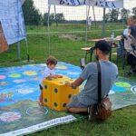 Pan klęczy z brzegu gry wielkoformatowej, obok stoi małe dziecko opierające się o kostkę do gry.