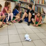 Dzieci siedzą na pufach i bawią się robotem.