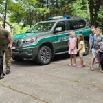 Dzieci stoją na tle samochodu straży granicznej z funkcjonariuszem jego psem.